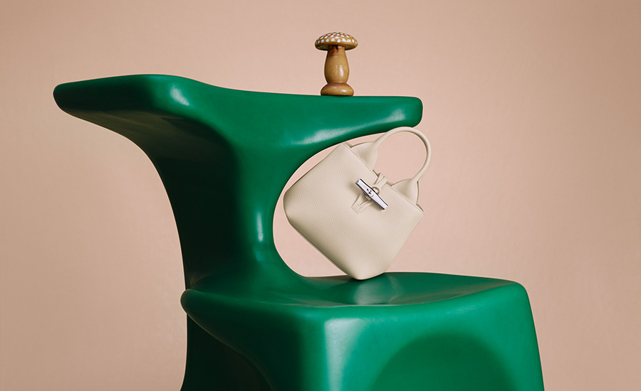 Die Le Rosseau Tasche in einem edlen Cremeweiß inszeniert für die Frühjahr-/Sommer 2025 Kampagne von Longchamp. Die Bag steht auf einem grünen, organisch geformten Stuhl, auf dessen Lehne ein kleiner Holzpilz platziert ist. Die Szenerie symbolisiert die enge Bindung der Maison zur Natur.)