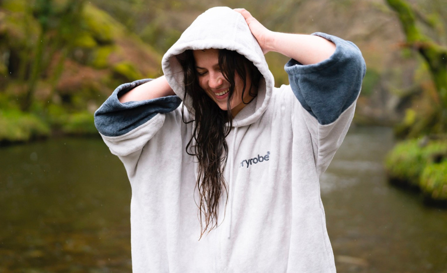 Dryrobe Towel Robe in hellem Grau – eine junge Frau mit nassem Haar und Kapuze lacht, während sie mitten in der Natur am Wasser steht.