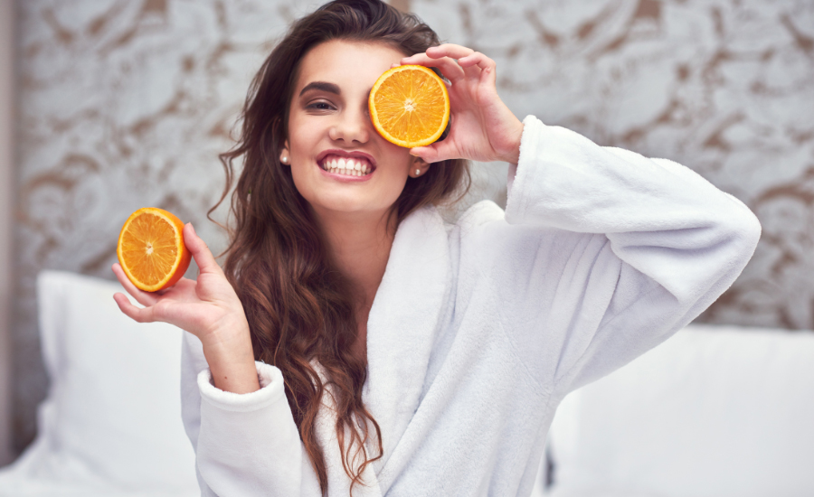 Altrient Vitamin C stärkt die Haut von innen; eine junge Frau hält zwei frisch aufgeschnittene Orangen vor Gesicht und Körper als Symbol für Vitamin-C-reiche Pflege und natürliche Ausstrahlung.
