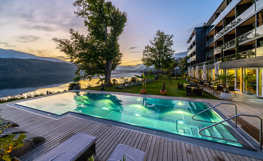 Beheizter Outdoor-Pool am See bei Sonnenuntergang, daneben ein modernes Wellnesshotel mit Terrasse und Liegestühlen.
