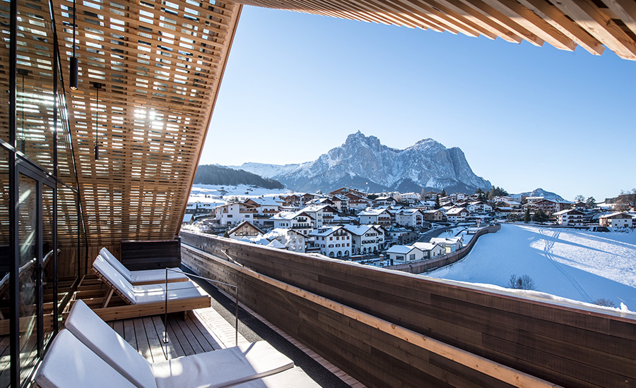 Sonnige Hotelterrasse mit Liegen und Holzfassade, Panoramablick auf ein verschneites Dorf und Berge im Hintergrund.