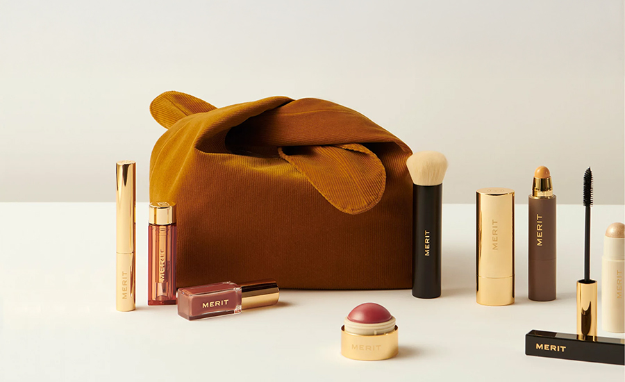 Ein Stillleben mit Merit Beauty Make-up und einer braunen Kosmetiktasche zeigt unter anderem Mascara, Lippenprodukt, Stick und Creme Blush auf hellem Hintergrund.