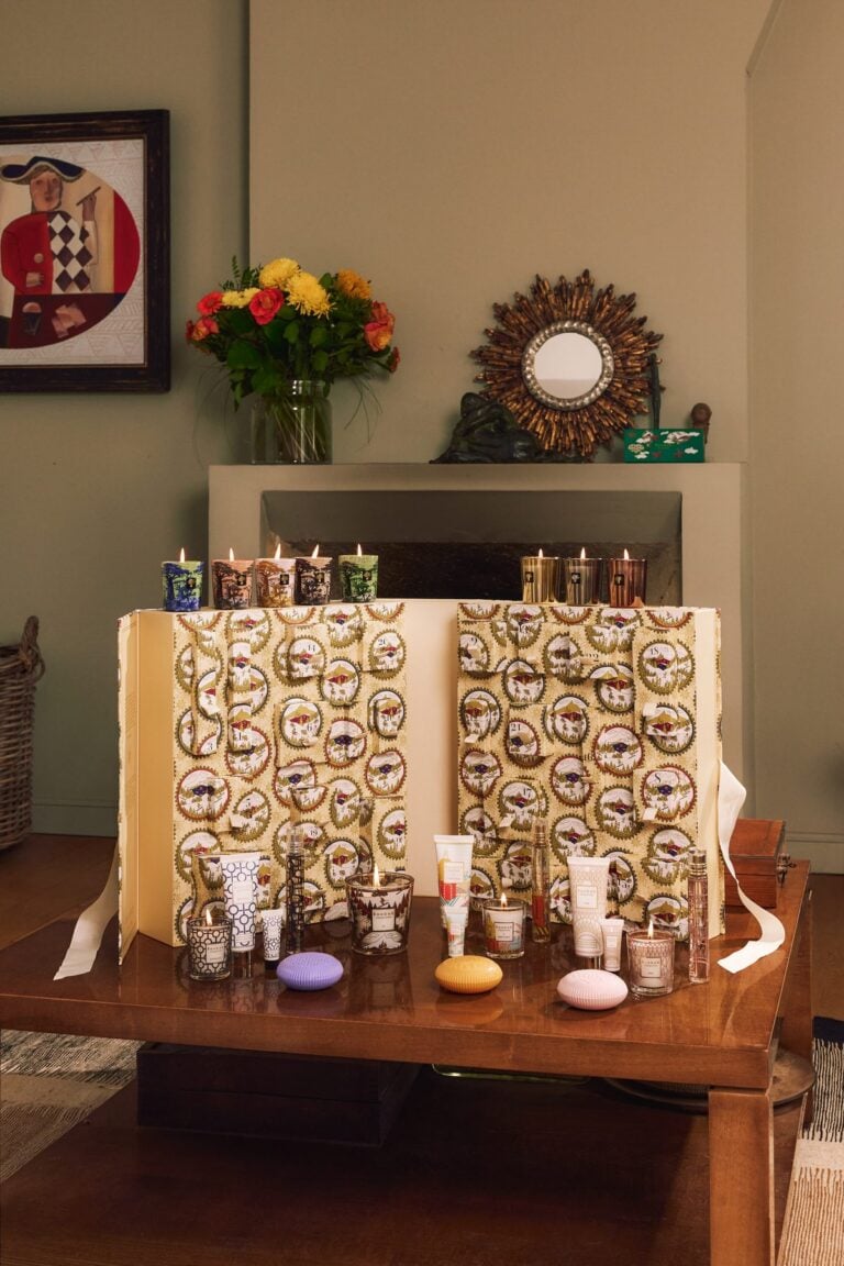 Aufgeklappter Adventskalender von Baobab Collection auf einem Holztisch vor einem Kamin, davor Duftkerzen, Seifen und Pflegeprodukte