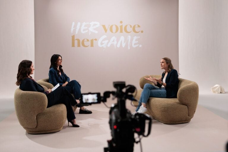 Drei Frauen sitzen in Sesseln im Studio und sprechen miteinander, im Vordergrund eine Kamera, an der Wand der Schriftzug „Her Voice, Her Game“