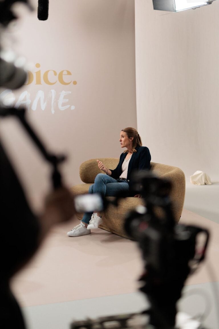 Justine Henin sitzt in einem hellen Sessel im Studio, im Vordergrund unscharf eine Kamera