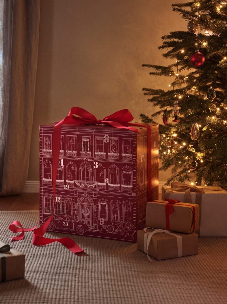 Roter Westwing-Adventskalender mit Schleife neben einem geschmückten Weihnachtsbaum und verpackten Geschenken